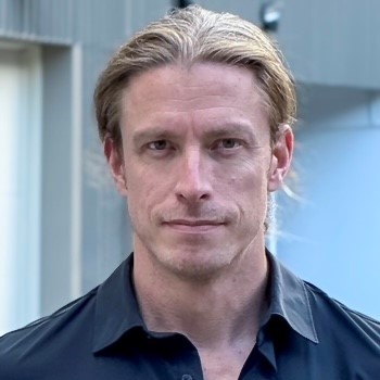 Head-and-shoulders portrait of a man with blond hair wearing a dark blue shirt, looking at the camera.